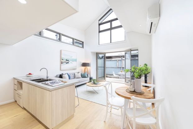 Bright open-plan living, dining and kitchen area with a light timber island, white cabinetry, pale timber flooring, compact sofa seating and a round dining table. The room has a high sloping ceiling, large clerestory and triangular windows, plus glazed doors opening to a small courtyard. The overall design is airy, contemporary and minimalist.