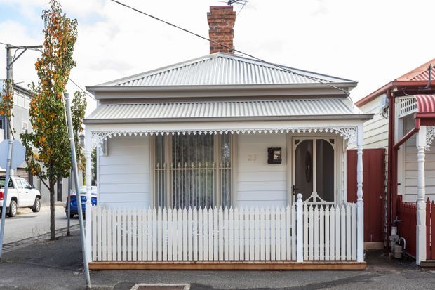 Street-level view of a compact single-storey cottage facade with white weatherboard cladding, a corrugated metal hipped roof, brick chimney, decorative lacework verandah trim, barred front window, and a small white picket fence. The house is closely surrounded by neighbouring urban dwellings.