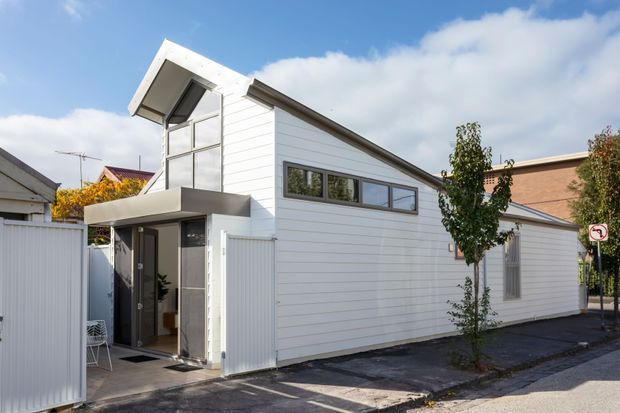 Exterior street view of a compact contemporary white-clad building with a sharply sloping roof, tall glazed entry volume, projecting flat canopy, and narrow horizontal windows. The building sits on a paved urban streetscape beside neighbouring homes, with a small planted tree and boundary fencing.