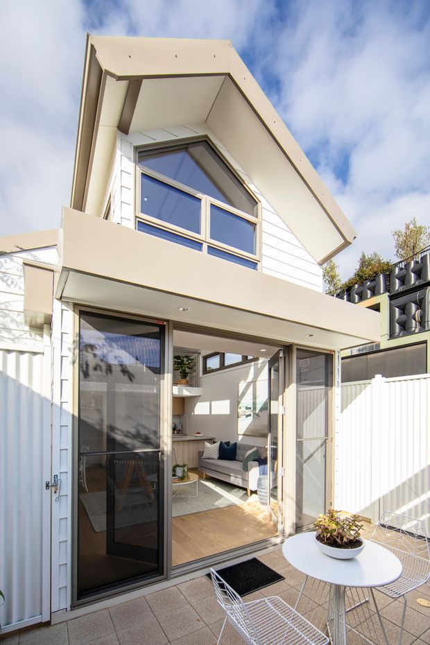 Bright exterior view of a compact multi-level home with a steep gable roof, white horizontal cladding, and a prominent upper-level window assembly. The ground floor opens onto a paved courtyard or patio through large glazed sliding doors, revealing a small living room with a sofa and timber flooring. Outdoor dining furniture occupies the foreground, while neighbouring structures and fencing frame the scene.