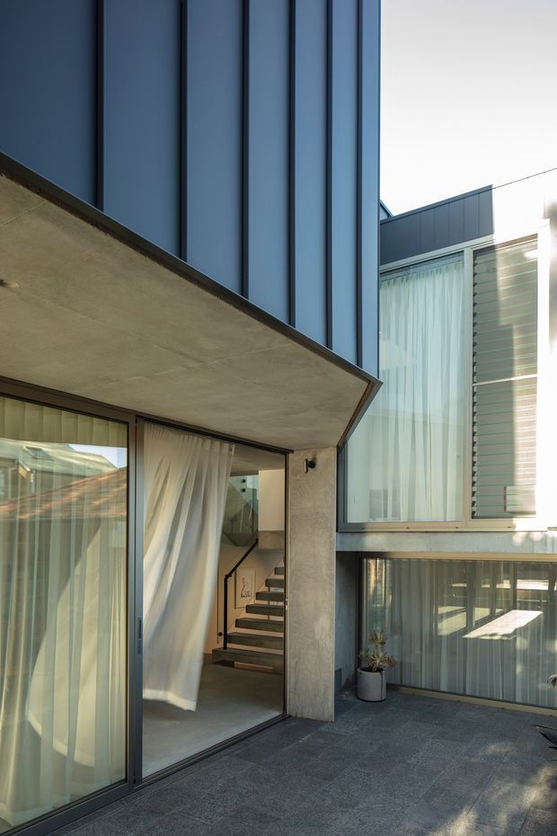 Exterior view of a contemporary multi-level residence focused on a recessed entry and courtyard-like terrace. The facade combines dark vertical metal cladding, smooth exposed concrete, extensive floor-to-ceiling glazing with sheer curtains, and horizontal light-toned cladding. A projecting concrete slab creates a strong cantilever over the glazed entrance, while an interior stair is visible through the open sliding glass door.