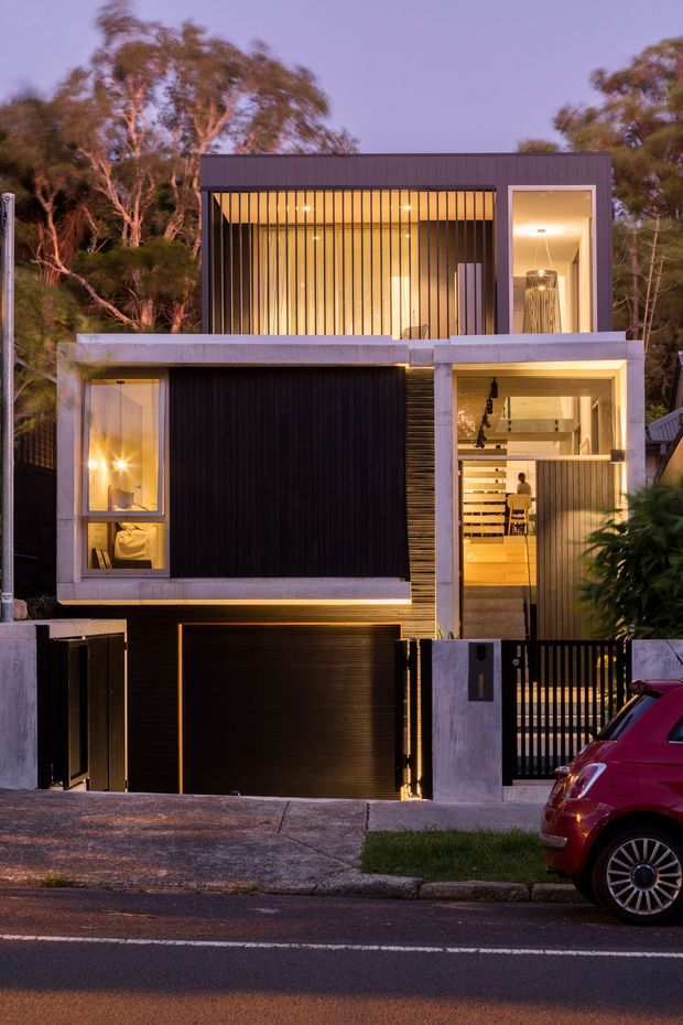 Twilight street-view photograph of a narrow, multi-level contemporary residence set among mature trees. The facade combines exposed pale concrete framing, dark vertical cladding and screening, extensive warm-lit glazing, a recessed garage, and a gated pedestrian entry. Vertical metal bars and a cantilevered upper level give the home a strongly geometric, private urban character.