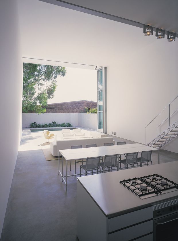 A bright, minimalist open-plan interior combining a kitchen, dining area and living space. The foreground features a large white island with an integrated gas cooktop and stainless-steel oven, while a long dining table with dark chairs occupies the centre. Beyond it, a white lounge arrangement opens through large folding glass doors to a paved courtyard with planting and a rendered boundary wall. A slender open staircase and exposed track lighting add contemporary architectural detail.