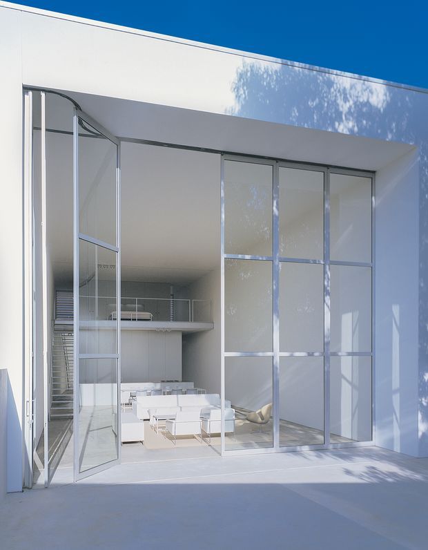 Exterior view of a stark white modernist building with a deep recessed opening and extensive floor-to-ceiling glazed doors and panels in slim aluminium frames. The glazing reveals a bright, minimalist living space with white low furniture, a compact kitchen or bar area, and a staircase. Strong geometric lines, crisp shadows, and a flat roof create a clean contemporary architectural composition.
