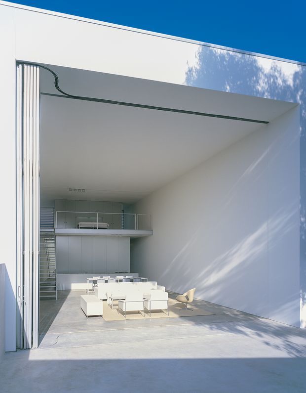 A minimalist white architectural interior/exterior threshold opens into a spacious double-height living area. The room features smooth white walls and ceiling, a polished light-grey concrete floor, floor-to-ceiling glazed folding or sliding doors, a mezzanine level with glass balustrade, a staircase, and sparse white modular seating with a sculptural tan chair. Strong sunlight and deep shadows emphasize the crisp modern geometry.