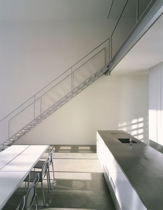 A bright, minimalist kitchen and dining area beneath an open stairway. The space features a long white dining table with slender metal chairs, a large white island with a dark grey benchtop and integrated sink, smooth white walls and cabinetry, polished concrete flooring, and a lightweight metal stair and balustrade. Strong sunlight enters from the right, casting geometric shadows across the floor and wall.