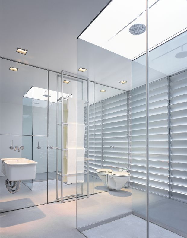 A bright, contemporary bathroom with a wall-mounted bathtub at left, a glass-enclosed shower, wall-hung toilets, extensive mirrored surfaces, and full-height horizontal louvre blinds along the right wall. The space has pale grey finishes, frameless glass partitions, recessed square ceiling lights, and a clean minimalist fit-out.