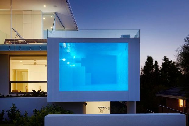 Twilight exterior view of a contemporary multi-level residence featuring a prominent illuminated swimming pool integrated into a projecting box-like structure. The home has crisp white geometric volumes, extensive glass balustrades and glazing, recessed lighting, and an open balcony or terrace. Dark landscaping and neighbouring roof forms frame the composition.