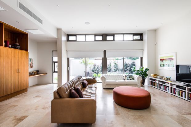 A bright, spacious contemporary living room with high ceilings and large glazed openings. The room features a long brown leather sofa, a cream upholstered sofa, a large rust-coloured round ottoman, pale stone-look flooring, timber built-in cabinetry and shelving, and a television and media unit. Clerestory windows and broad sliding glass doors bring in abundant natural light, with a landscaped courtyard visible beyond.