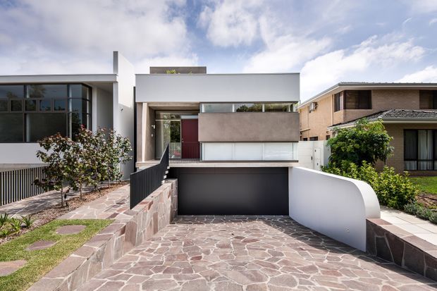 Street-facing view of a contemporary multi-storey residence with a flat roof, box-like upper-level form, recessed entry, dark garage opening, and a curved white privacy wall. The frontage combines pale rendered surfaces, extensive glazing, dark metal balustrades, stone retaining walls, and irregular natural-stone driveway paving, with neighbouring homes visible on either side.