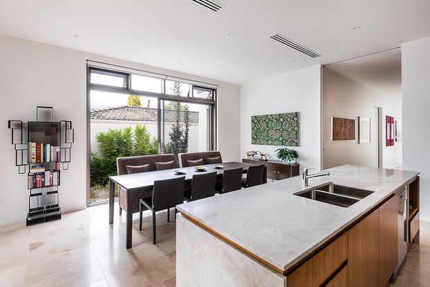Bright open-plan kitchen and dining area with a large stone island and integrated sink in the foreground. A dark timber dining table with upholstered chairs sits beside expansive windows, while wall-mounted shelving, artwork, and timber cabinetry add detail. The space has a clean, contemporary fit-out with abundant natural light and a connection to a planted courtyard.