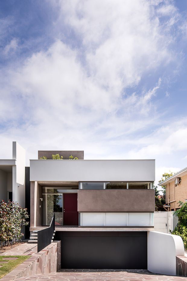 Front elevation of a narrow multi-storey urban residence with a minimalist contemporary facade. The house features crisp white rendered walls, a projecting upper-level volume with a muted grey-brown finish, dark glazing and garage door, a deep red entry door, and a curved white boundary wall. A paved driveway and stepped front path lead to the street, with neighbouring homes and landscaping visible at the edges.