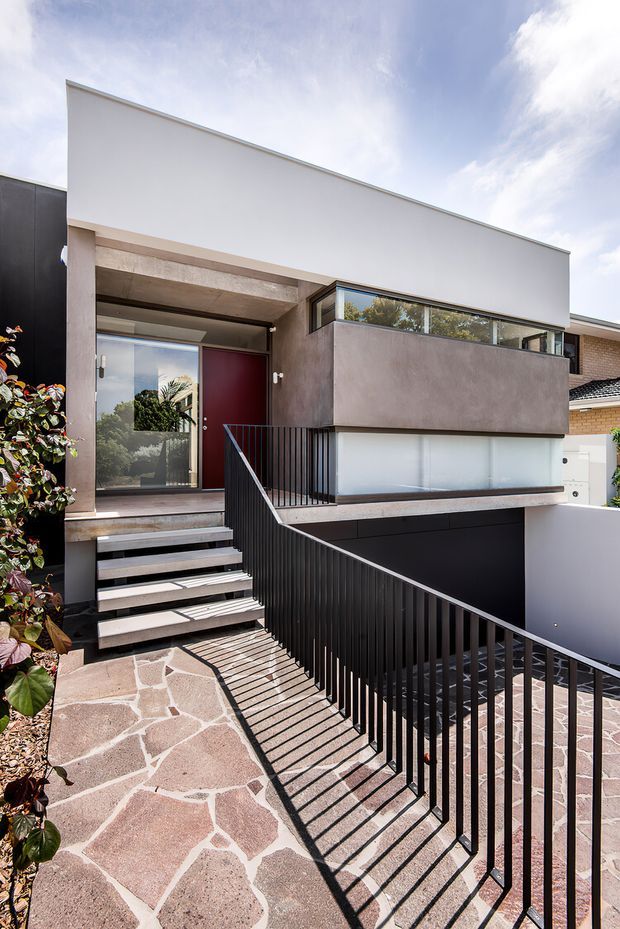 Photograph of a contemporary multi-level residential facade viewed from the front entry and driveway. The house features a crisp white upper volume, dark charcoal side wall and garage door, projecting rendered balcony or bedroom volume, exposed concrete entry canopy, large glazing, a deep red front door, black metal balustrades, and irregular natural stone paving. The sloped approach and lower-level garage create a strong split-level composition.