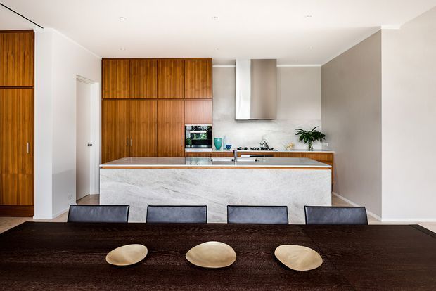 A contemporary open-plan kitchen and dining area viewed from the dining table. The kitchen features a large marble-look island with an integrated cooktop, white walls and cabinetry accents, a stainless-steel rangehood, and extensive warm timber veneer cabinetry with vertical and horizontal panelling. A dark timber dining table with four dark upholstered chairs and sculptural bowls occupies the foreground.