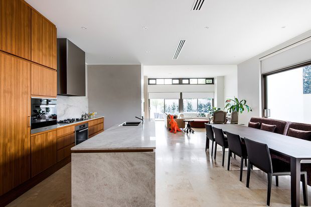 Bright open-plan interior combining a contemporary kitchen, dining area and living space. The kitchen features tall warm timber cabinetry, integrated appliances, a long pale stone island and a dark stone or marble-look splashback. A large dining table with dark chairs occupies the foreground, while extensive glazing and a high-level clerestory bring natural light into the room. The overall aesthetic is refined, minimal and warm, with polished pale flooring and clean white ceilings.
