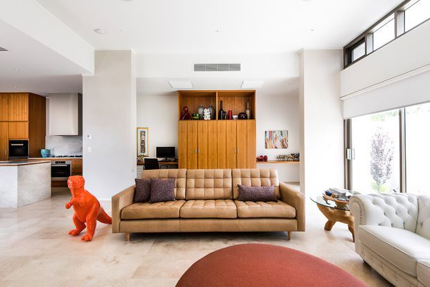 Bright, spacious open-plan living room with a tan tufted sofa, white sculptural armchair, rounded red ottoman, and a large timber built-in cabinetry and shelving wall. The kitchen is visible to the left, while tall glazing and high clerestory windows bring in abundant daylight. The space combines warm timber joinery, pale stone-like flooring, neutral walls, and playful sculptural decor.