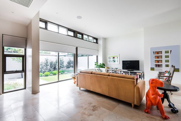 Bright, spacious contemporary living room with a large tan leather sectional sofa, television and low media joinery, polished pale stone or concrete-look flooring, and extensive floor-to-ceiling glazing. High-level clerestory windows and a linear ceiling vent bring in abundant daylight, while a vivid orange sculptural chair adds a playful accent. The glazing opens visually to a landscaped garden.