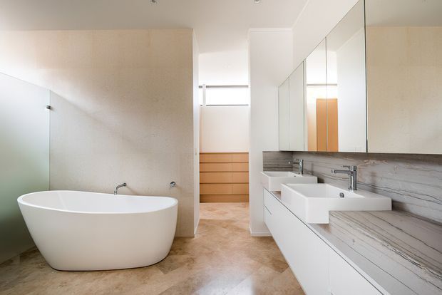 Bright contemporary bathroom with a freestanding white bathtub, double vanity, wall-to-wall mirrored cabinetry, and a long grey-veined stone benchtop and backsplash. Warm beige stone-look flooring and a timber drawer unit visible through the opening add contrast to the predominantly white and light neutral scheme.