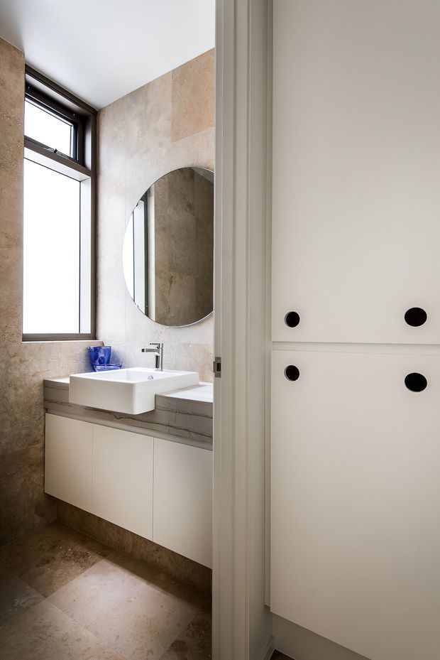 A refined bathroom viewed through a partially open doorway, featuring a wall-mounted white vessel sink on a pale stone vanity, matching stone wall and floor surfaces, a large round mirror, dark-framed window, and white built-in cabinetry with black circular pulls. The room has a clean, contemporary feel with warm neutral finishes and natural light.