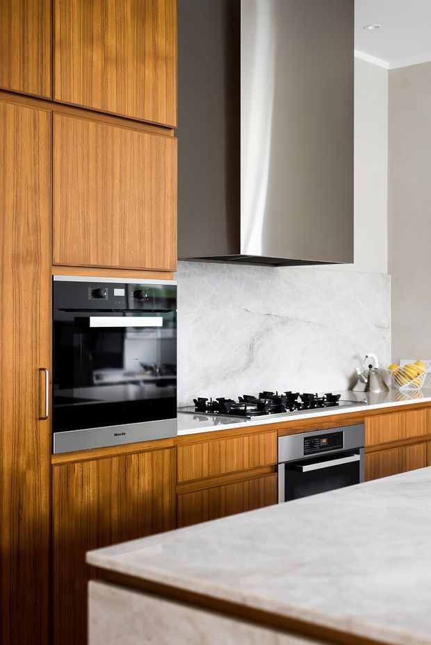 A refined kitchen featuring warm vertical-grain timber cabinetry, integrated black and stainless-steel appliances, a gas cooktop, a pale veined stone splashback, and a large matching stone island in the foreground. The cabinetry extends nearly to the ceiling, while recessed ceiling lights and neutral plaster walls create a clean, contemporary composition.