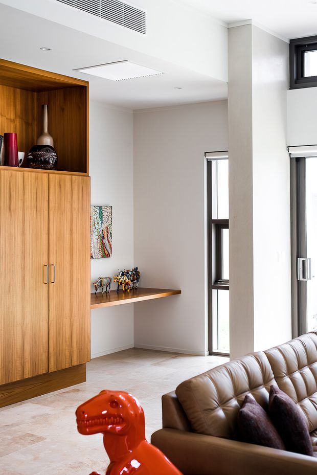 Bright contemporary living room interior with a tall built-in timber display cabinet and shelving, pale walls and stone-look flooring. A tufted brown leather sofa occupies the foreground, with a glossy red dinosaur sculpture and decorative objects adding playful accents. Tall black-framed windows and a prominent structural column bring strong vertical lines and natural light into the space.