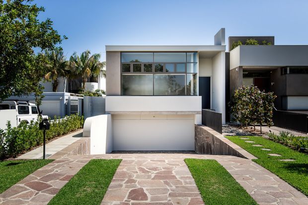 Street-facing view of a compact modern residence with a flat-roofed, white rendered facade, broad upper-level glazing, an integrated garage, and a paved driveway with grass strips. Tropical planting and neighbouring contemporary homes frame the property.