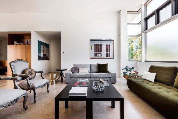 Bright, spacious contemporary living room with very high ceilings and large black-framed windows. The room features a grey sofa, olive-green sofas, ornate upholstered armchairs, a large black rectangular coffee table, pale flooring, white walls, and built-in timber-toned joinery. A mezzanine-like upper window band and clerestory glazing bring in abundant natural light.