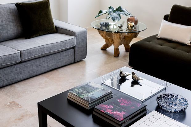 A refined living room with a grey upholstered sofa and black lounge chair arranged around a black rectangular coffee table. The table displays stacked books, decorative birds, a blue-and-white bowl, and a white architectural model or sculptural object. A round glass side table with an ornate sculptural wooden base sits between the seating, with a potted plant and decorative objects. The room has pale stone or tile flooring, light walls, and a restrained neutral palette with dark accents.