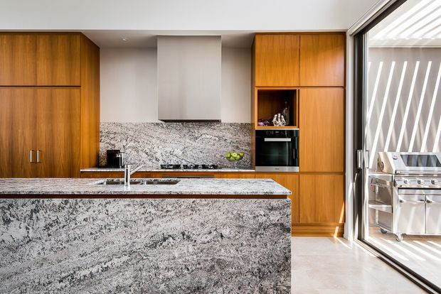 A bright, contemporary kitchen with extensive warm timber cabinetry, a large stone island with integrated sink and seating overhang, matching stone splashback, stainless-steel appliances, and full-height glazing opening toward an outdoor barbecue area. The compact, highly streamlined layout has strong natural light and a clean material palette.