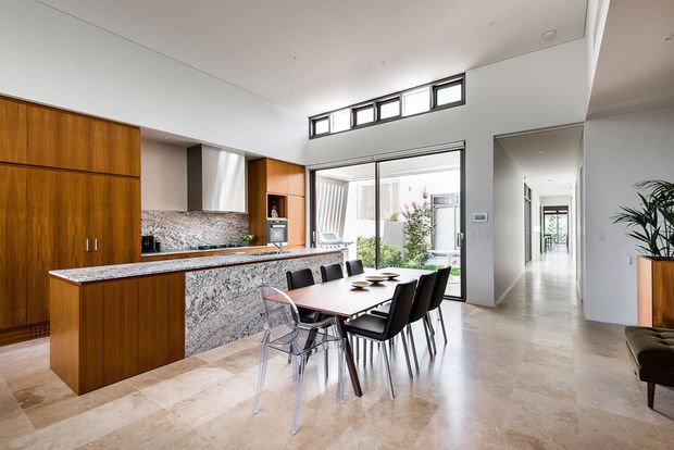 Bright open-plan kitchen and dining area with warm timber cabinetry, a dramatic patterned stone island and splashback, a modern dining table with mixed seating, and pale stone flooring. High clerestory windows and large glazed doors bring in abundant daylight, while a hallway and indoor courtyard or garden are visible beyond.