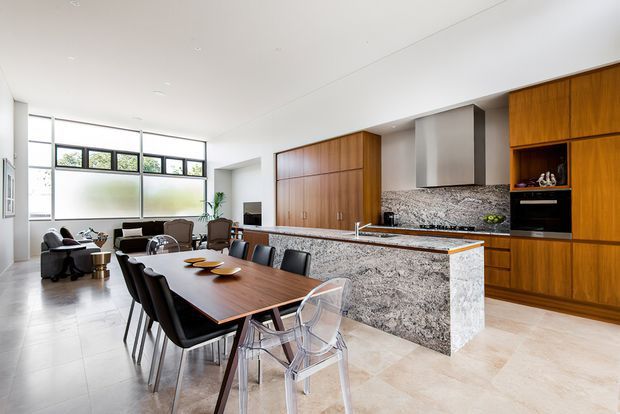 Open-plan contemporary kitchen and dining area with a large rectangular timber dining table, mixed black and transparent dining chairs, and a lounge seating zone beyond. The kitchen features extensive warm timber cabinetry, a grey-speckled natural-stone island and backsplash, integrated appliances, and a clean white ceiling with expansive glazing and clerestory windows. The space is bright, spacious, and visually connected to the living area.