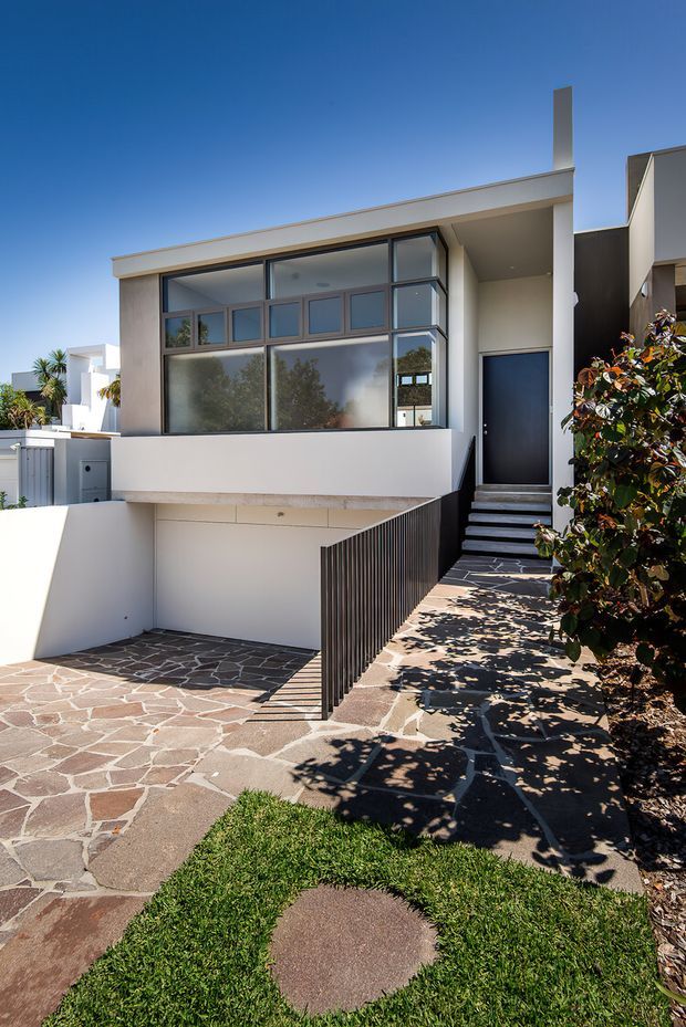 Street-facing view of a contemporary multi-level residence with a flat roof, pale rendered walls, extensive dark-framed glazing, a recessed entry, and a narrow stairway behind a dark vertical-bar balustrade. The forecourt uses irregular flagstone paving with curved lawn inserts and landscaped planting.