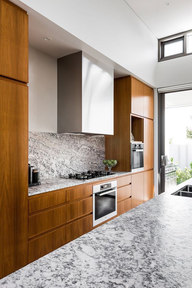 Bright contemporary kitchen with extensive warm timber cabinetry, a dramatic grey-and-white veined stone benchtop and matching splashback, integrated stainless-steel appliances, gas cooktop, and a large island. White walls and ceiling, dark-framed windows, and an open glazed door provide abundant natural light. The space has a tall ceiling and clean built-in joinery.