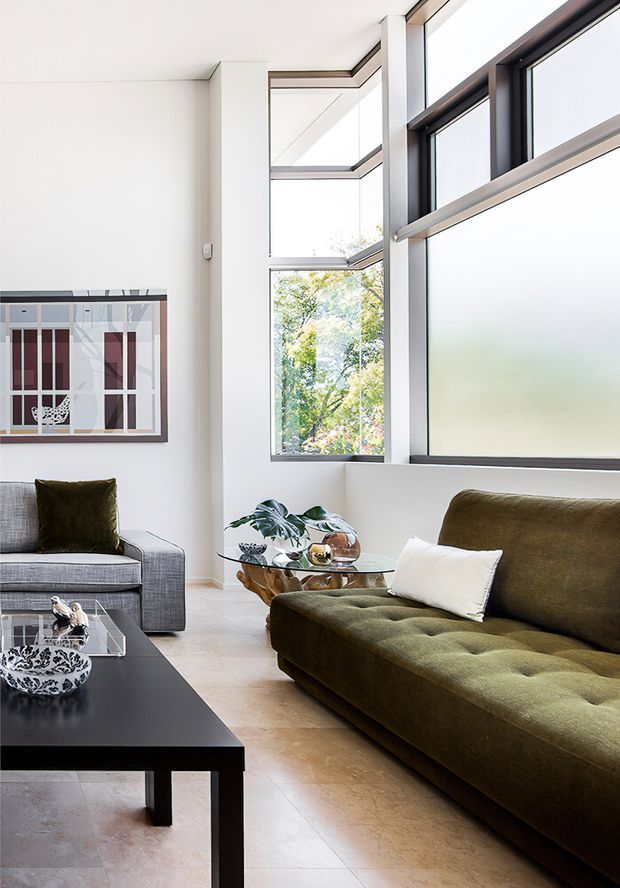 A bright, refined living room with large black-framed windows, pale walls and flooring, a deep olive-green tufted sofa, a grey upholstered armchair, and a black rectangular coffee table. A glass side table with decorative objects and plants sits near the windows, while a framed abstract artwork adds visual interest. The space has abundant natural light and a clean, tailored contemporary feel.