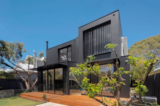 Exterior view of a contemporary two-storey residential home with a sculptural dark facade, extensive glazing, vertical screening, and a warm timber deck. The building sits within a landscaped suburban setting under a clear blue sky.