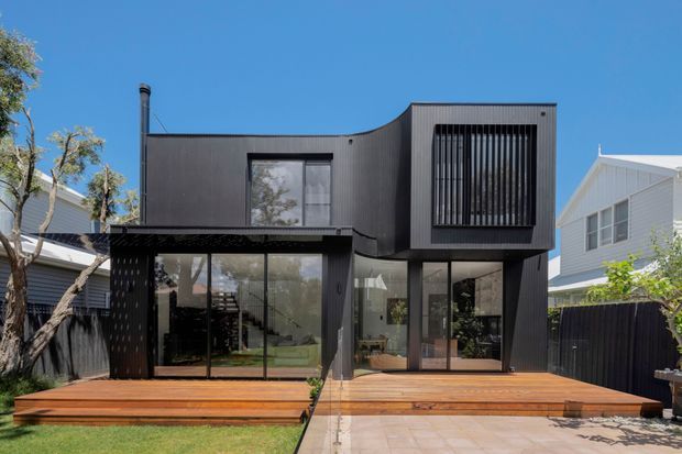 Contemporary two-storey dark-clad house with a projecting upper volume, large glazed ground-floor openings, and a broad timber deck and paved outdoor area. The facade features vertical ribbed cladding, a prominent black vertical-slat window screen, and a curved roofline transition. Neighbouring homes and landscaping frame the residential exterior.