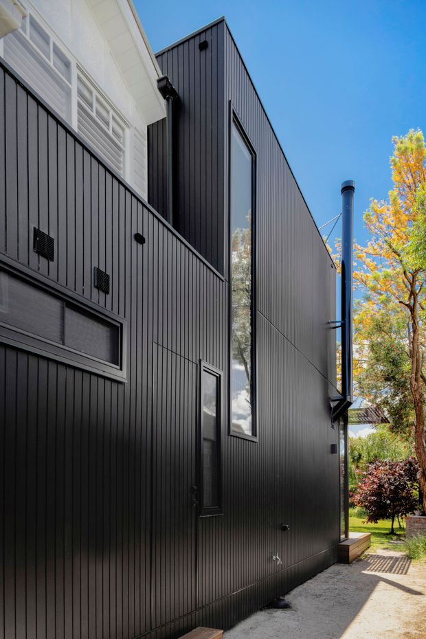 Exterior view of a tall, narrow modern residential extension clad in dark vertically ribbed metal or composite panels. The facade features slim vertical windows, a projecting black flue or chimney, and a contrasting white upper section with horizontal windows. A narrow paved walkway runs alongside the building, with established trees and garden planting visible beyond. Bright daylight and clear blue sky emphasize the strong black-and-white facade contrast.