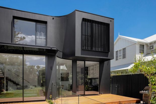 Exterior view of a contemporary two-storey home extension with dark vertically ribbed cladding, expansive floor-to-ceiling glazing, a projecting upper volume with vertical screening, and a timber deck. A lighter weatherboard house is visible adjacent in the background.