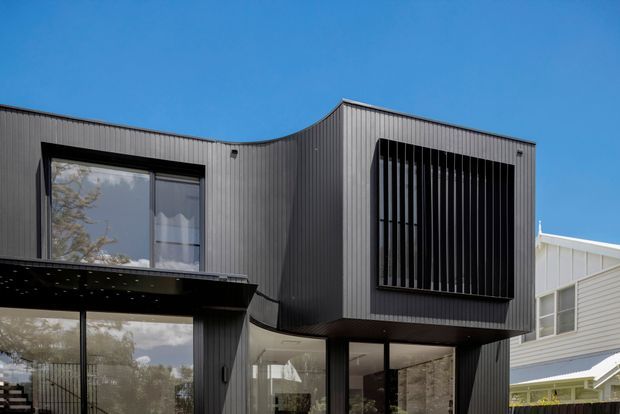 Exterior view of a contemporary two-storey residence with a sculptural curved roofline, dark vertically ribbed metal cladding, large glazed openings, and a prominent screened upper-level window. A lighter neighbouring building is visible in the background under a clear blue sky.