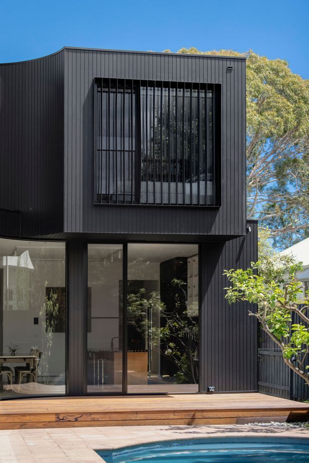 A contemporary two-storey residential extension or rear facade clad in dark vertical metal or fibre-cement-style panels. Large black-framed glazed doors and windows open onto a timber deck beside a swimming pool. The upper-level window has prominent vertical security bars or screening, while mature trees and neighbouring residential elements are visible around the building.