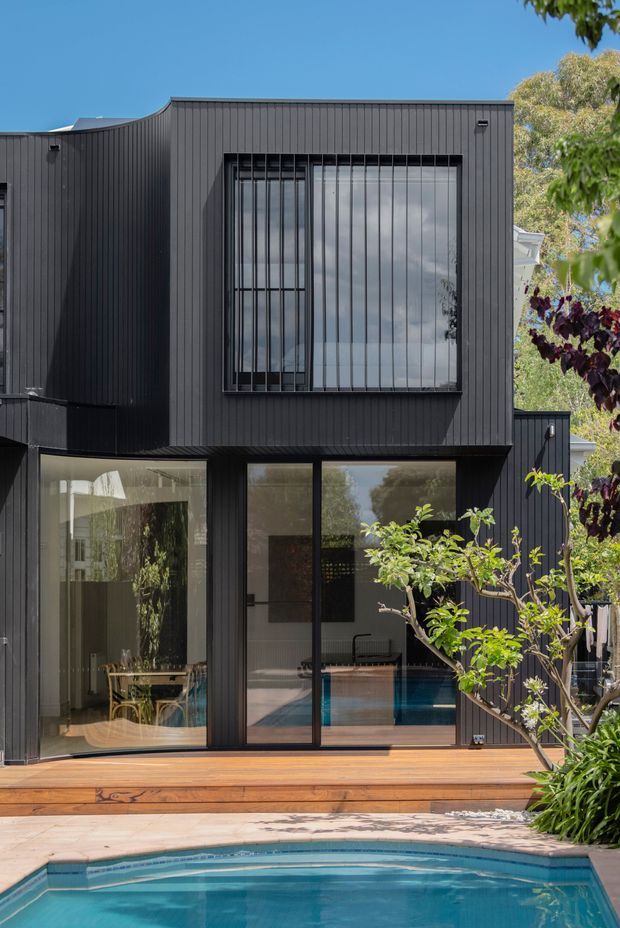 A contemporary two-storey dark-clad home viewed from a landscaped pool area. The facade features vertically ribbed black cladding, large black-framed glazed doors and windows, and a projecting upper level with a barred-looking window screen. A timber deck borders the swimming pool, with mature planting around the edges. Interior dining and living areas are visible through the glazing.