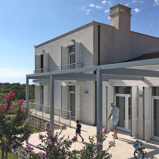 Exterior view of a bright, multi-level rendered masonry house with a flat roof, chimney, balconies, pale grey metal pergola structure, shuttered windows and extensive paved outdoor terraces. Mediterranean-style planting and a stone retaining wall surround the property.