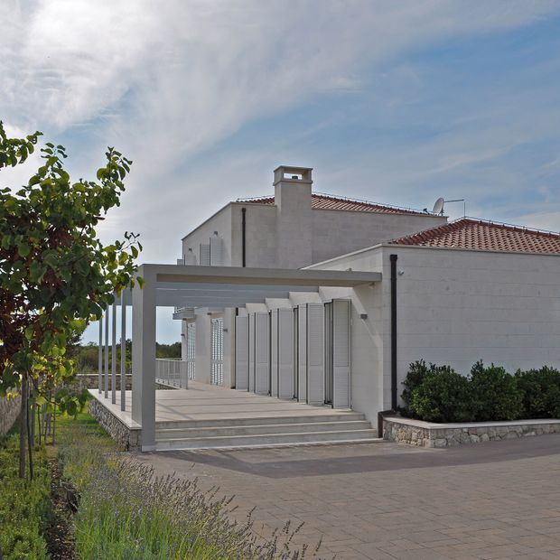 Exterior view of a white rendered masonry house with a terracotta tiled roof, chimney, covered entry terrace, broad steps, shutters, slender columns, and low stone-edged landscaping. The design has a clean Mediterranean influence and is photographed from the driveway or front approach.