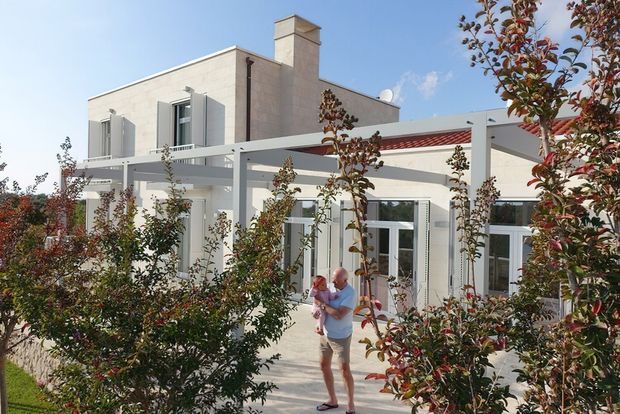 Exterior view of a contemporary light-coloured masonry house with a flat roof, prominent chimney, large glazed doors, and a white pergola or covered outdoor structure. Young landscaped trees and shrubs frame the paved outdoor area, with people present near the entrance. The architecture has clean geometric lines and a bright, open appearance.