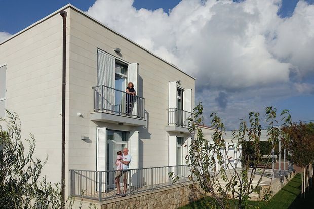 Exterior view of a contemporary two-storey residential building with pale horizontal cladding, white-framed glazed doors and windows, and narrow metal-railed balconies. A stone retaining wall and landscaped garden sit alongside the building, with people standing on the balconies. The structure has a simple pitched roof and a clean, modern facade under a bright partly cloudy sky.