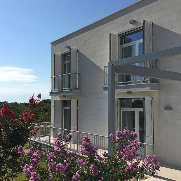 Exterior view of a contemporary multi-level residence with a pale rendered or masonry facade, tall windows, external louvred shutters, slim metal balcony railings, and a projecting grey structural or shading frame. Flowering shrubs occupy the foreground, with a clear blue sky and distant landscape beyond.