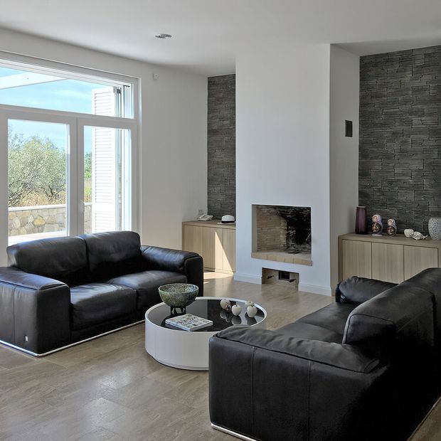 Contemporary living room with two black leather sofas, a round white coffee table, light timber flooring, and a central fireplace. White walls frame dark stacked-stone feature surfaces around the fireplace and built-in cabinetry. Large glazed doors provide abundant daylight and views to greenery outside.