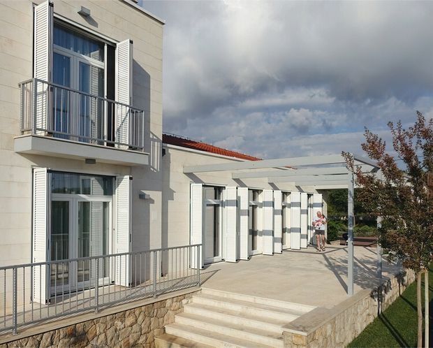 Exterior view of a light-coloured multi-level building with white louvred shutters, glazed balcony doors, metal balustrades, a broad paved terrace and pergola-like overhead structure. Stone retaining walls and steps lead up to the terrace, with a red tiled roof section and landscaped edge visible under a dramatic cloudy sky.
