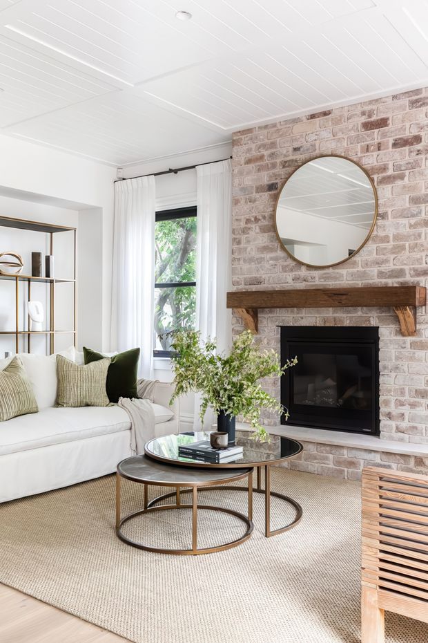 Bright, inviting living room with a white slipcovered sofa, layered neutral cushions, nested round coffee tables, open shelving, and a black fireplace set into a whitewashed exposed-brick feature wall. A timber mantel, round brass-framed mirror, white panelled ceiling, tall window with sheer curtains, and textured neutral rug create a warm contemporary-traditional interior.