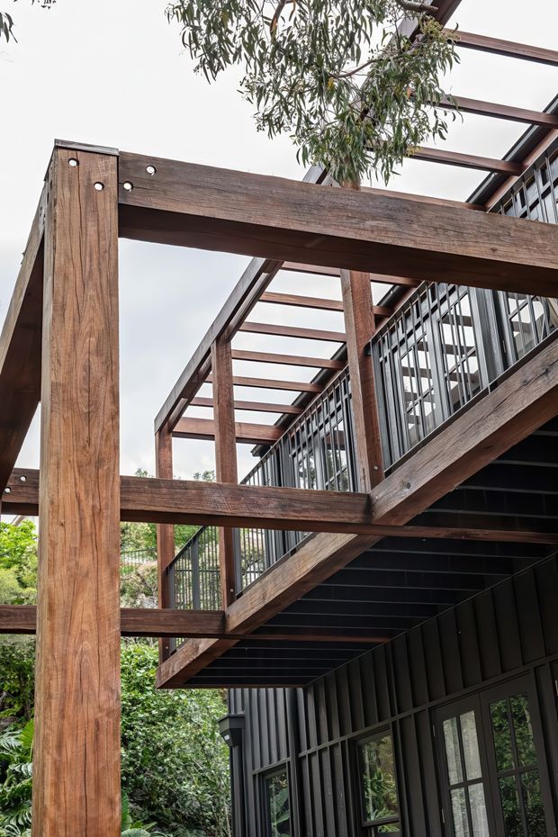 A close-up architectural view of a multi-level exterior deck and stair structure. Massive weathered timber posts and beams form an open pergola-like frame, with dark metal or timber cladding, black-framed glazing, vertical balustrades, and exposed dark stair treads. Dense vegetation is visible around the building, and a tree rises through the open overhead structure.